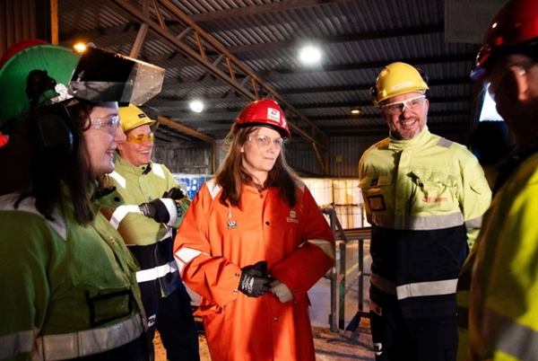 En gruppe personer i verneutstyr står samlet inne i et industrilokale og snakker sammen.