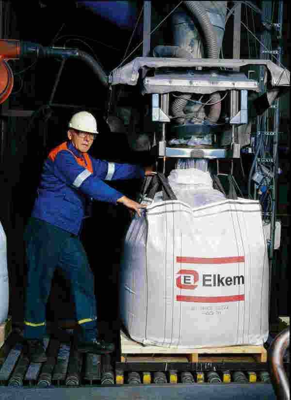 Historisk bilde av arbeider med sikkerhetshjelm håndterer en stor Elkem-sekk i industrielt miljø.